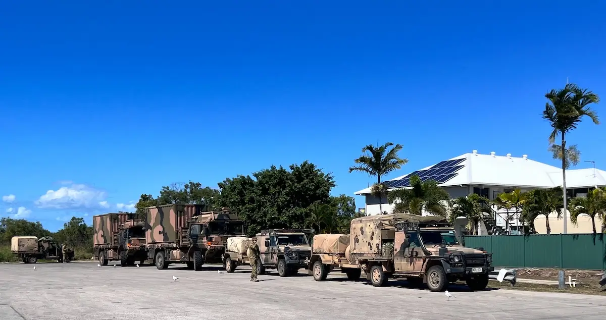 A line of military vehicles with camouflaged patterns is parked on a paved area. Soldiers stand near the trucks. In the background, there are palm trees and a modern white building with solar panels under a clear blue sky.