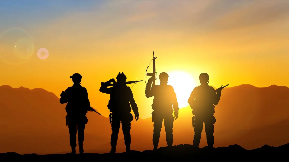 Silhouetted against a glowing sunset, four soldiers stand on a ridge. Each holds a weapon, with one saluting. The background features mountains and a brilliant orange sky.